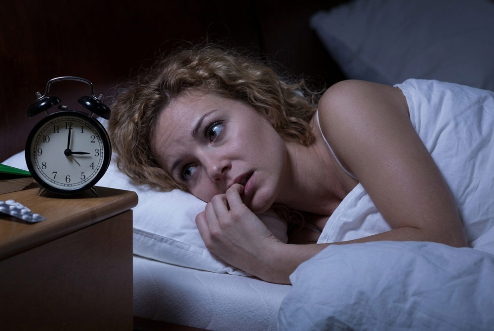 Anxious woman lying awake in bed at night looking at an alarm clock, illustrating sleep problems caused by stress and physical tension