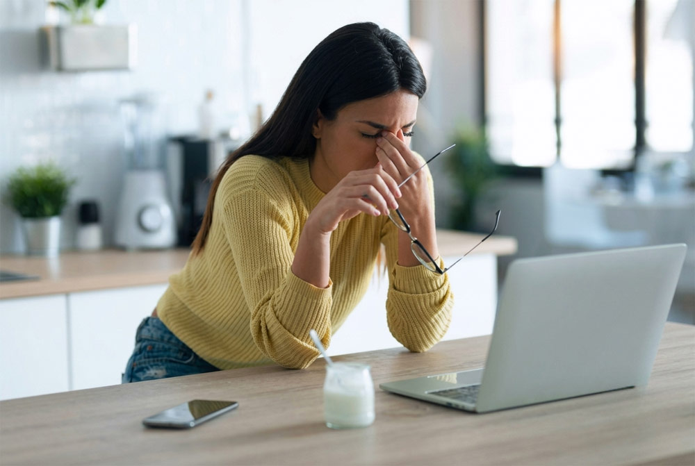 stressed woman working from home Woman taking off glasses and rubbing her nose bridge in stress while working on a laptop at home, illustrating work-from-home tension relieved by mobile massage in York