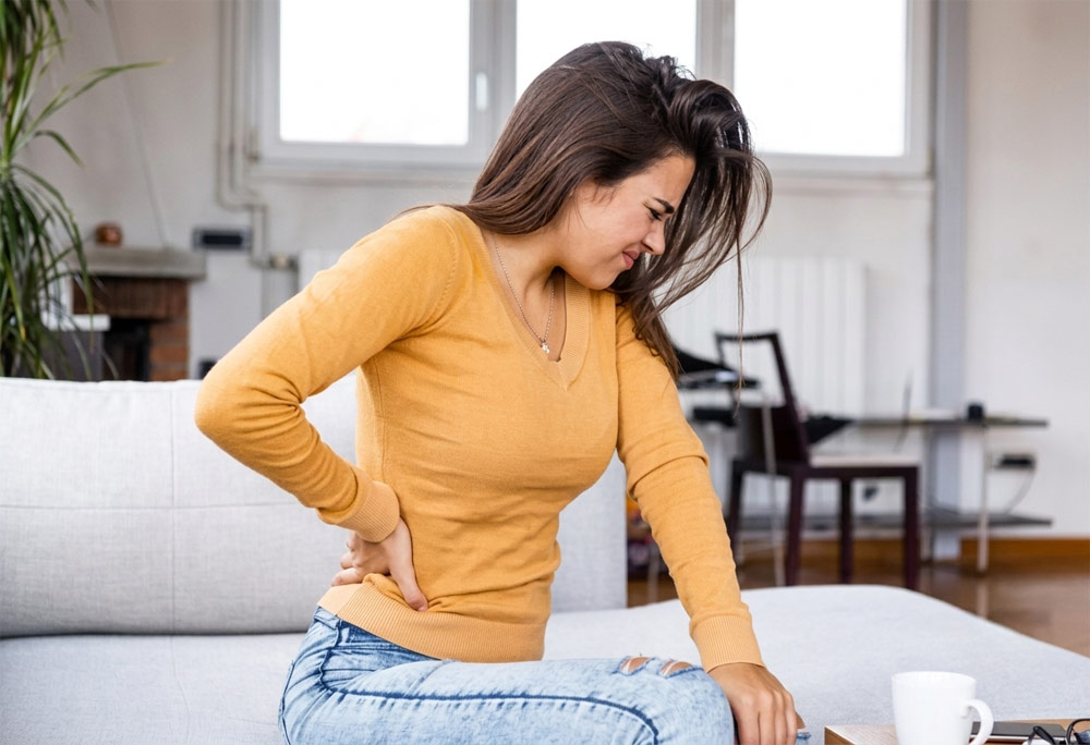 woman with bad back needing massage at home Woman sitting on her sofa at home holding her lower back in pain, illustrating the need for targeted mobile sports and deep tissue massage in York