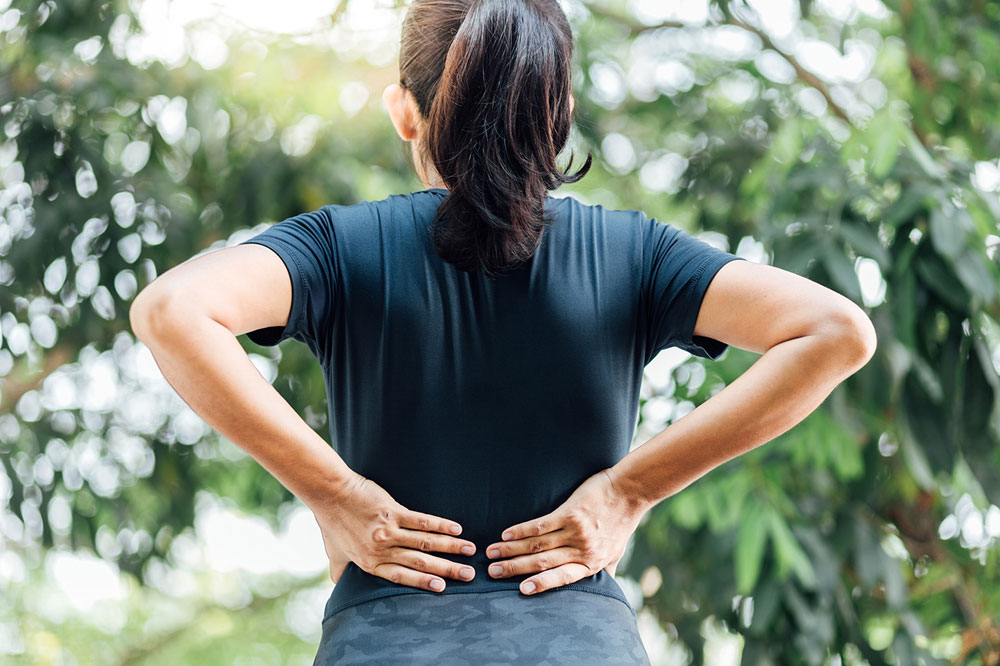 Woman in athletic wear holding her lower back in pain outdoors, illustrating back injury triggers from jogging or weight training addressed by a York sports massage therapist.
