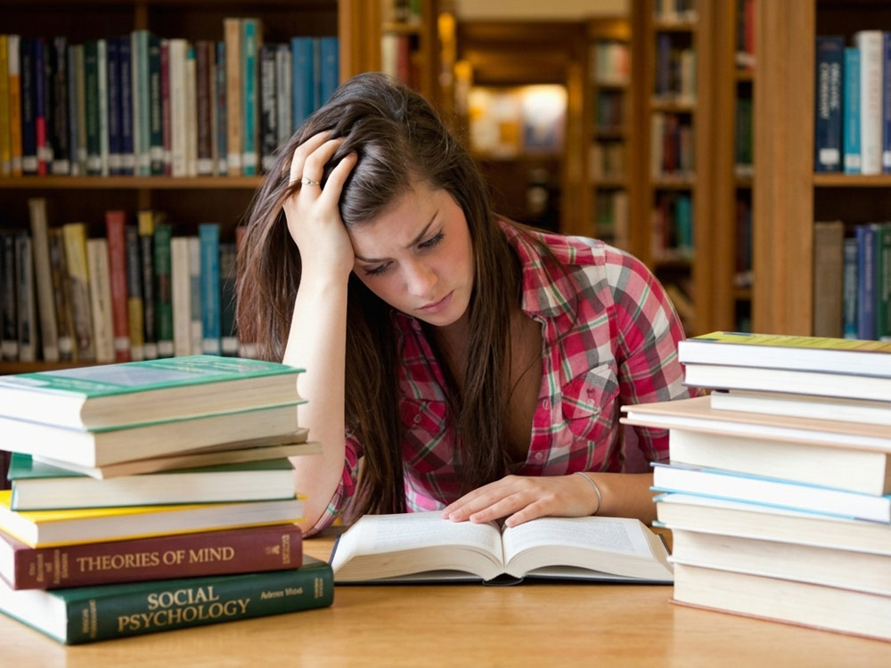 Stressed student holding her head while studying in a library, illustrating the academic pressure and neck strain solved by mobile massage in York